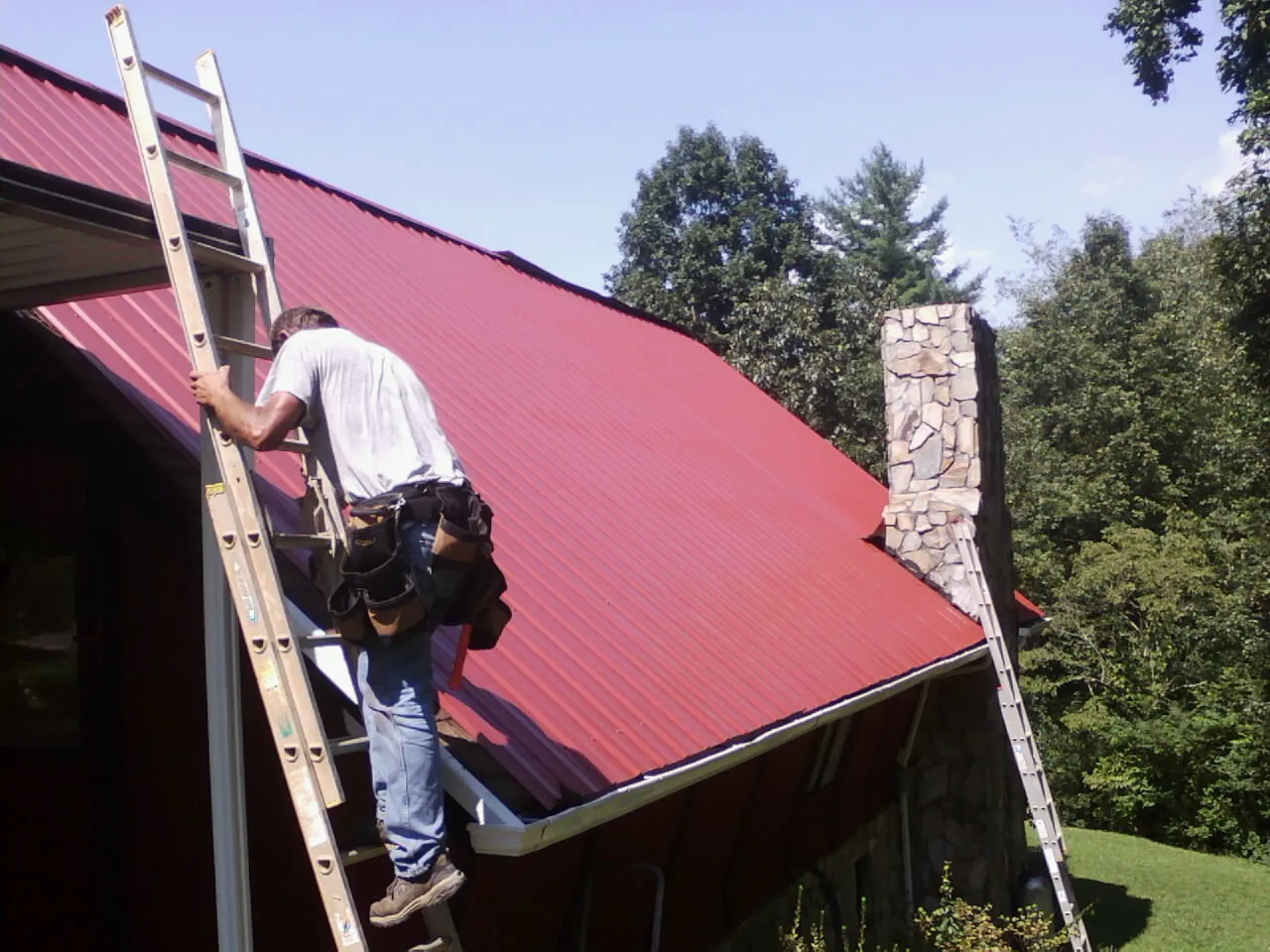 Roofer on a ladder beside a red metal roof and stone chimney — Soffit & Fascia Repair by Roofing Brothers in Naugatuck